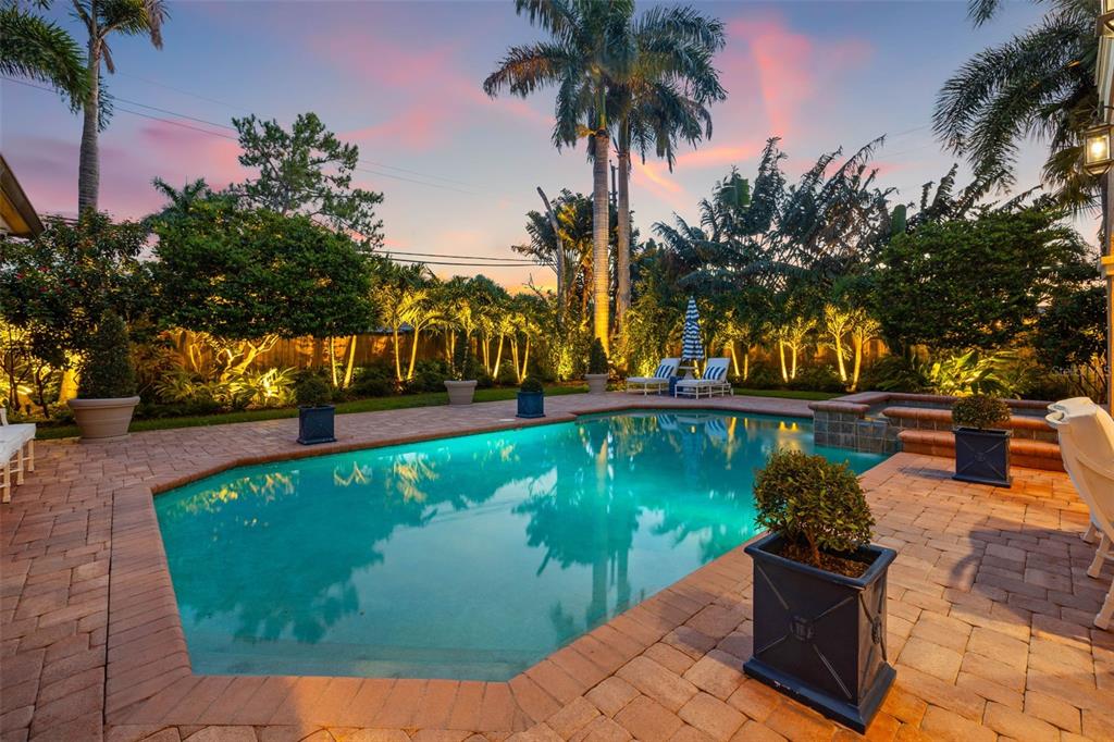 A backyard swimming pool with a stone patio, potted plants, and palm trees at sunset — 1124 SEVILLE LANE NE, ST PETERSBURG, FL