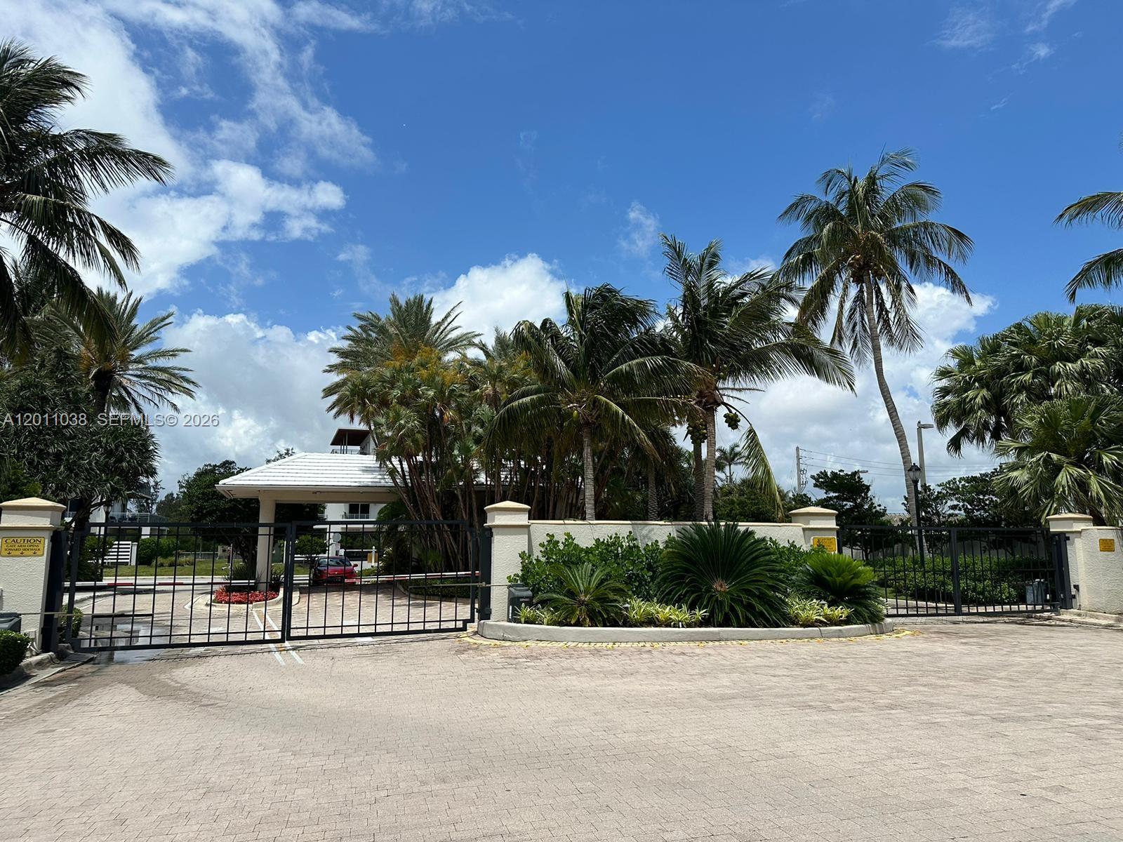 A gated entrance with a paved driveway, lush landscaping, and palm trees under a blue sky — 6001 N Ocean Dr # 503, Hollywood FL 33019, HOLLYWOOD, FL