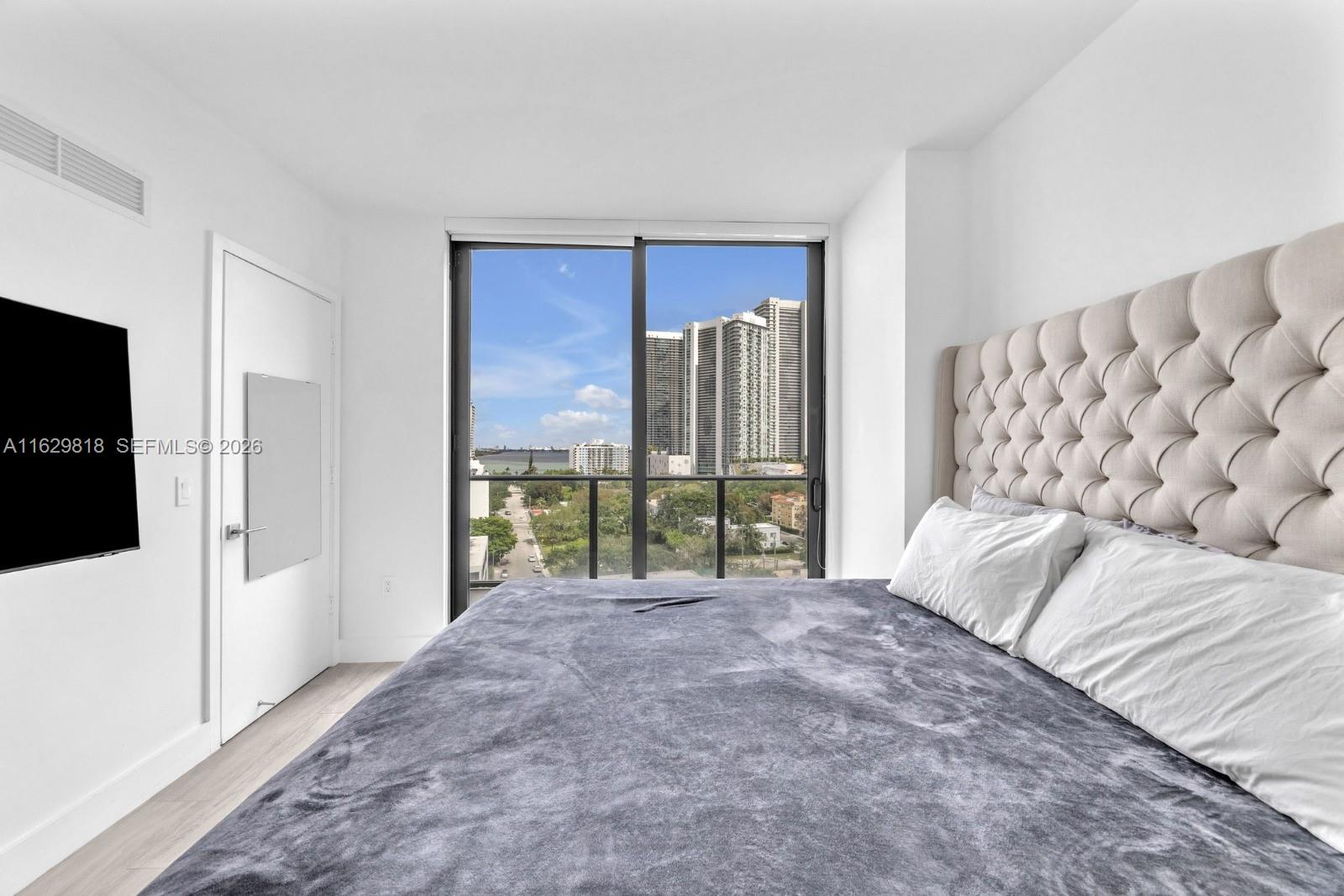 A bedroom features a tufted headboard, a gray bedspread, and a window overlooking city buildings — 121 NE 34th St # 905, Miami FL 33137, MIAMI, FL