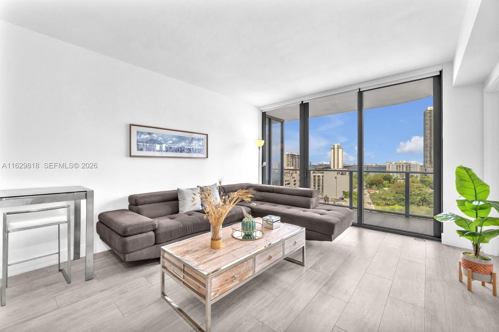 A modern living room features a gray sectional sofa, wooden coffee table, and floor-to-ceiling windows — 121 NE 34th St # 905, Miami FL 33137, MIAMI, FL