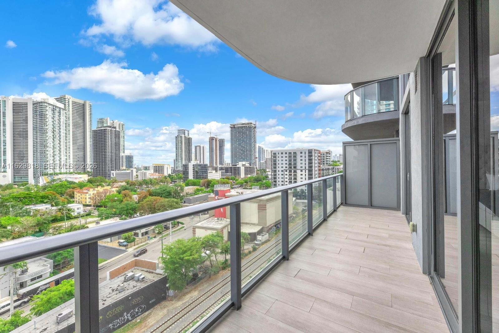 A modern apartment balcony features light wood-look tile flooring, a glass railing, and city views — 121 NE 34th St # 905, Miami FL 33137, MIAMI, FL