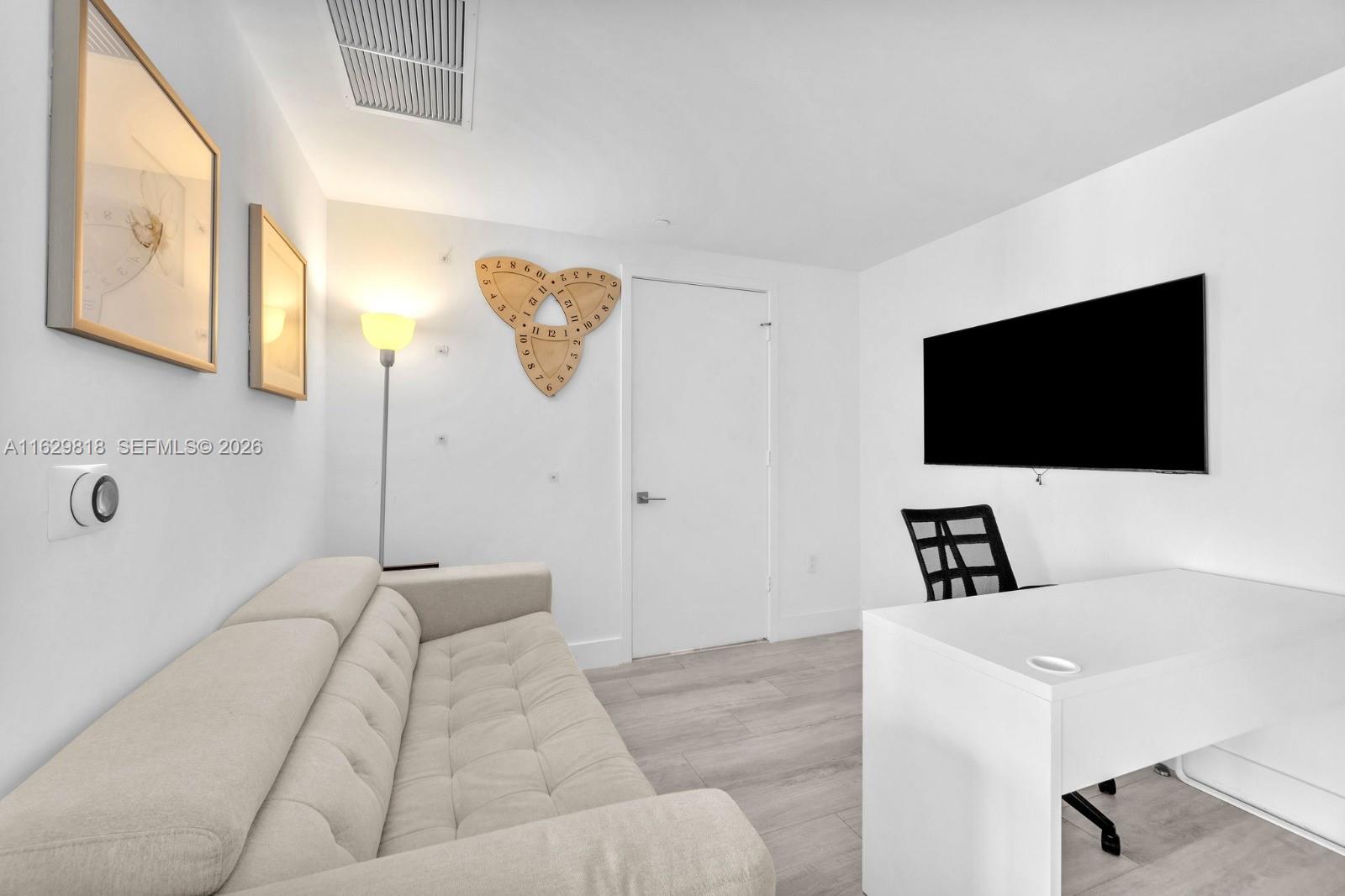 A room features a beige sofa, a white desk, a wall-mounted television, and a unique clock — 121 NE 34th St # 905, Miami FL 33137, MIAMI, FL