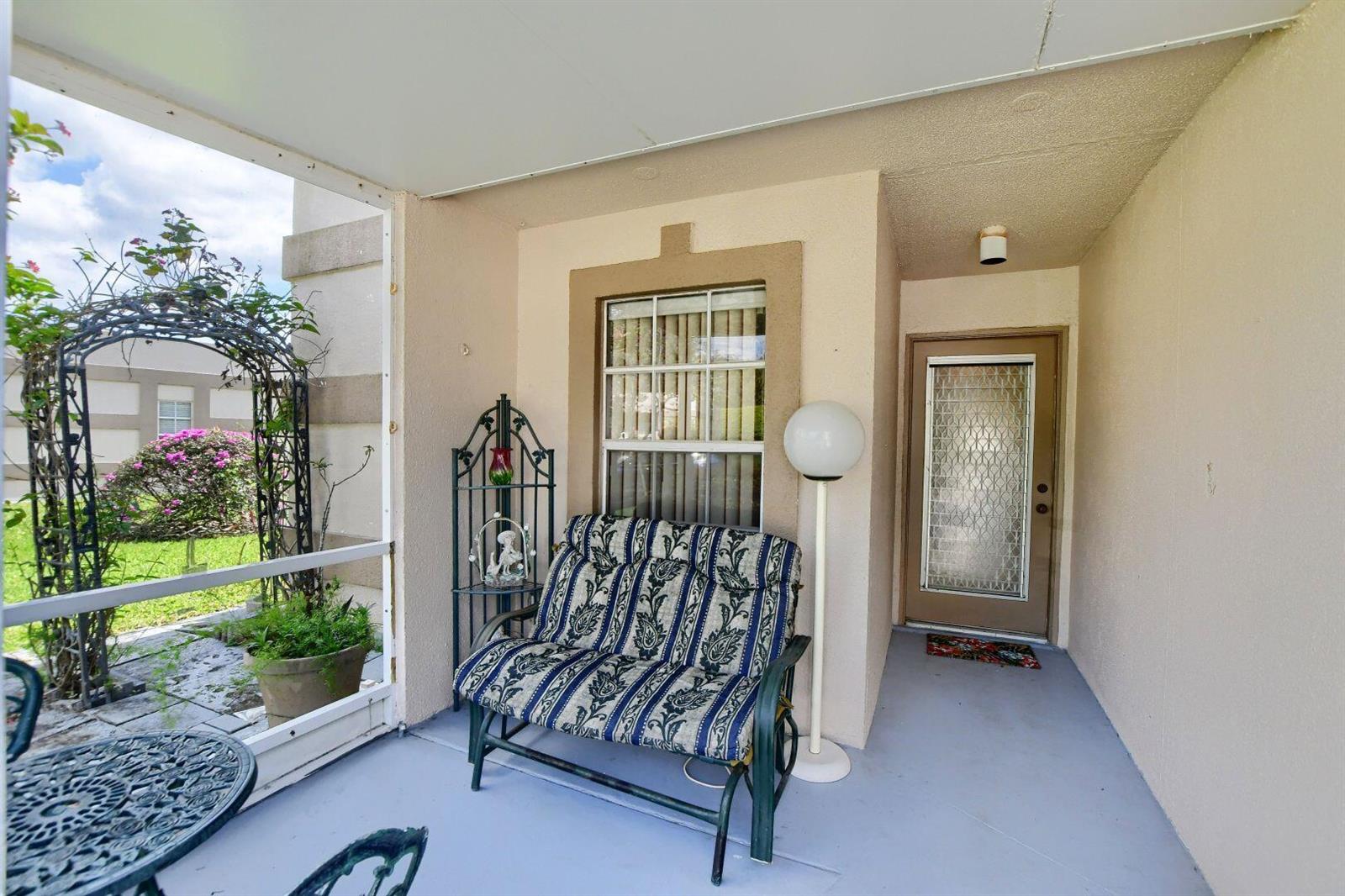 A covered patio features a patterned loveseat, a floor lamp, and a glass-paneled entry door — 9244 Flynn Circle # 4, Boca Raton FL 33496, BOCA RATON, FL