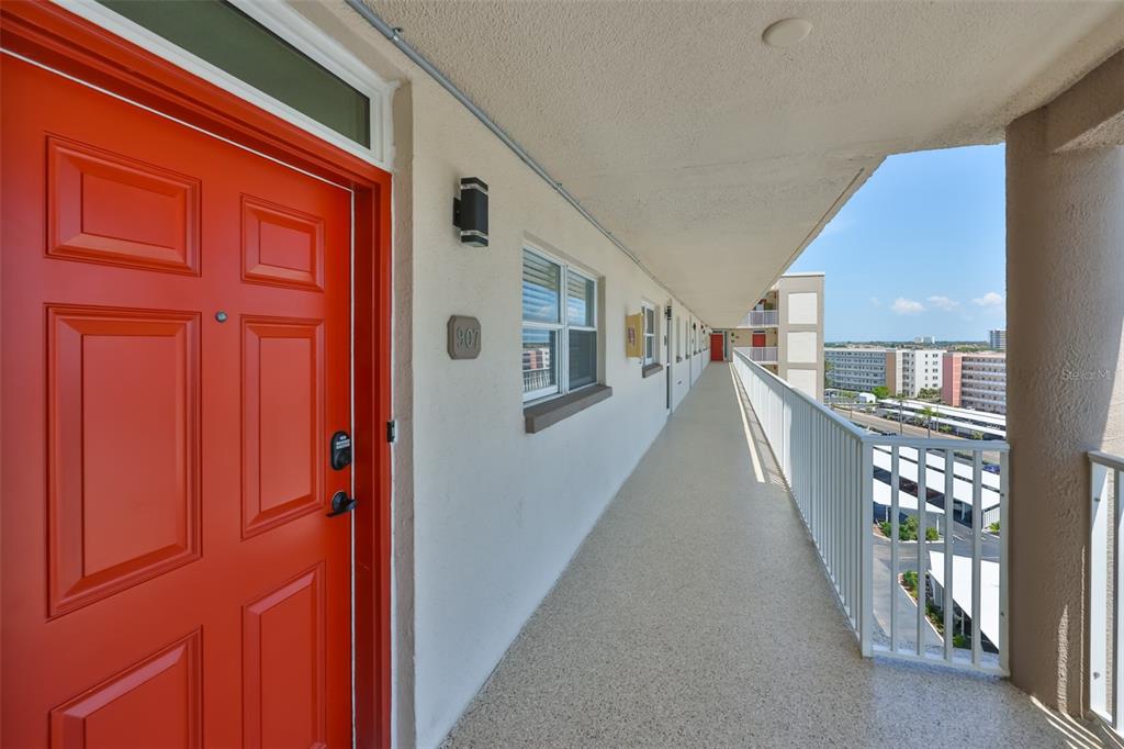 An exterior apartment hallway features a bright orange door, white railing, and concrete flooring — 5980 SHORE BOULEVARD S UNIT 907, GULFPORT, FL