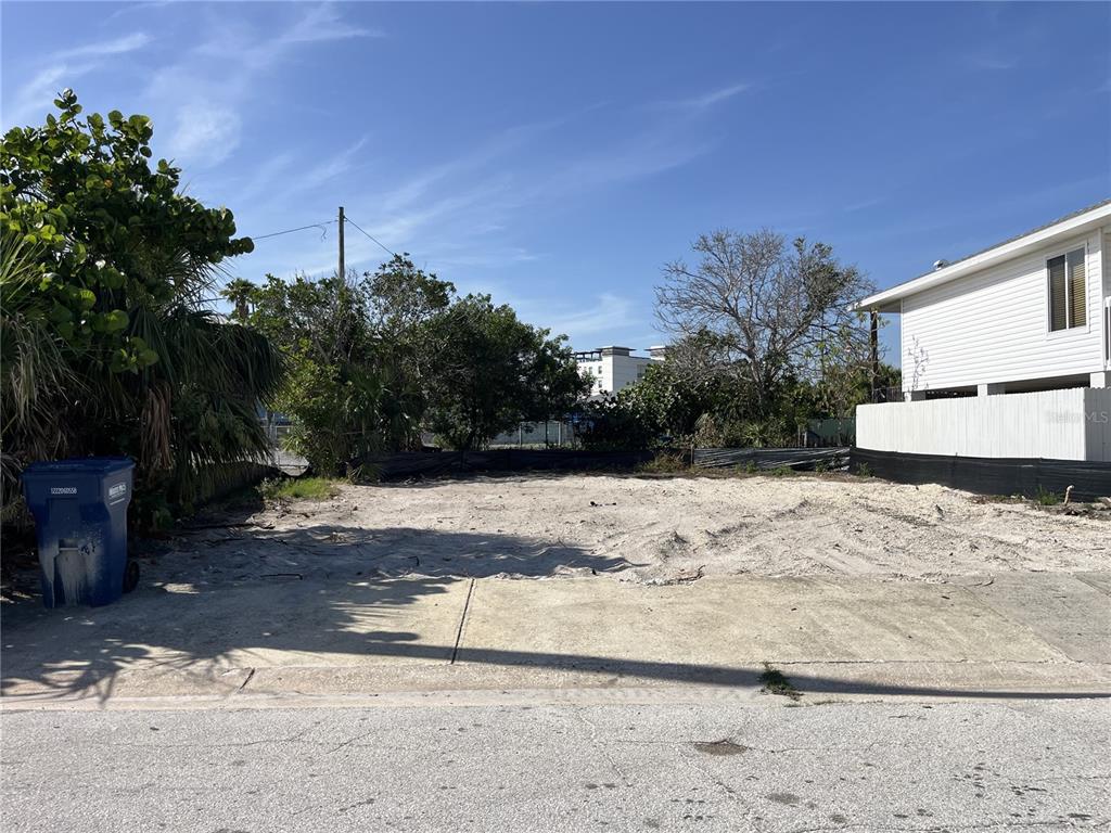 A vacant, sandy residential lot sits between a blue trash bin and a white house — 180 E MADEIRA AVENUE, MADEIRA BEACH, FL