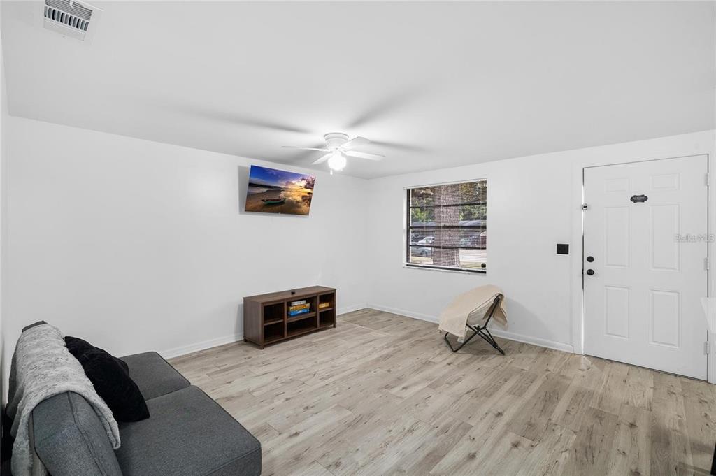 A living room with light wood-look flooring, a gray sofa, a television, and a white door — 1220 E 142ND AVENUE UNIT A, TAMPA, FL