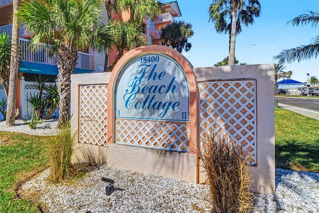 A beige stucco sign for The Beach Cottage stands in a landscaped yard with palms — 18400 GULF BOULEVARD UNIT 1302, INDIAN SHORES, FL
