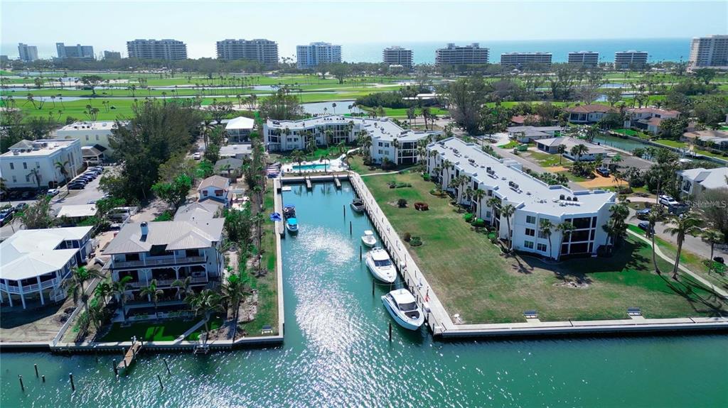 An aerial view shows a canal with docked boats between residential buildings and a golf course — 450 GULF OF MEXICO DRIVE UNIT 303, LONGBOAT KEY, FL