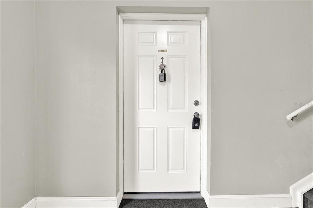 A white door with a lockbox and a peephole is centered on a grey wall — 9130 TUDOR DRIVE UNIT M105, TAMPA, FL