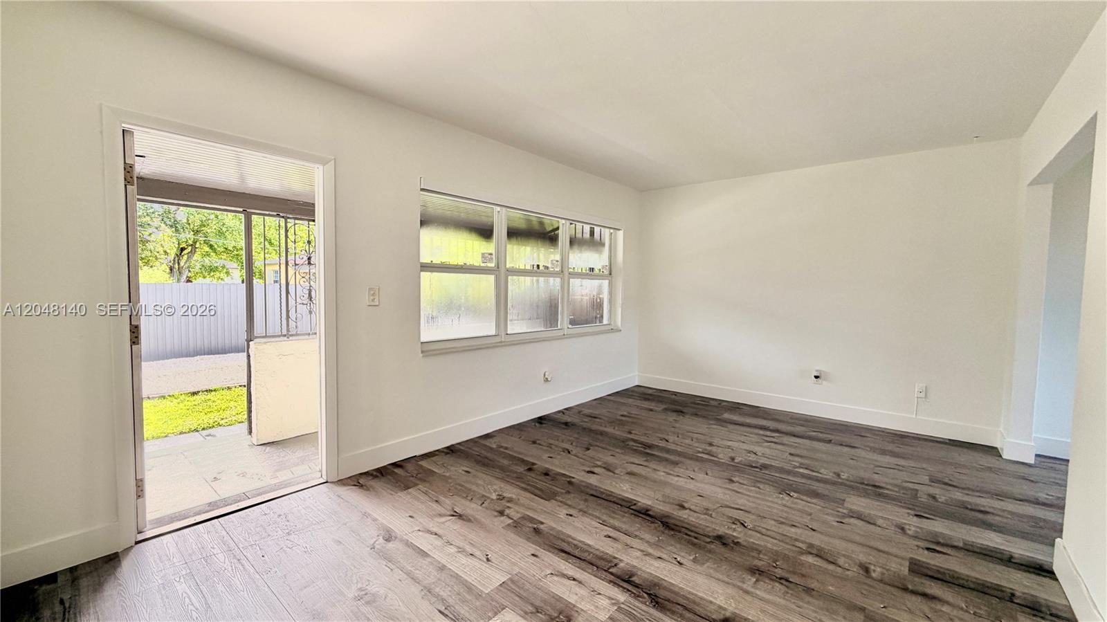 An empty room features white walls, dark wood-look flooring, a large window, and a door — 115 NW 118th St # 115, Miami FL 33168, MIAMI, FL