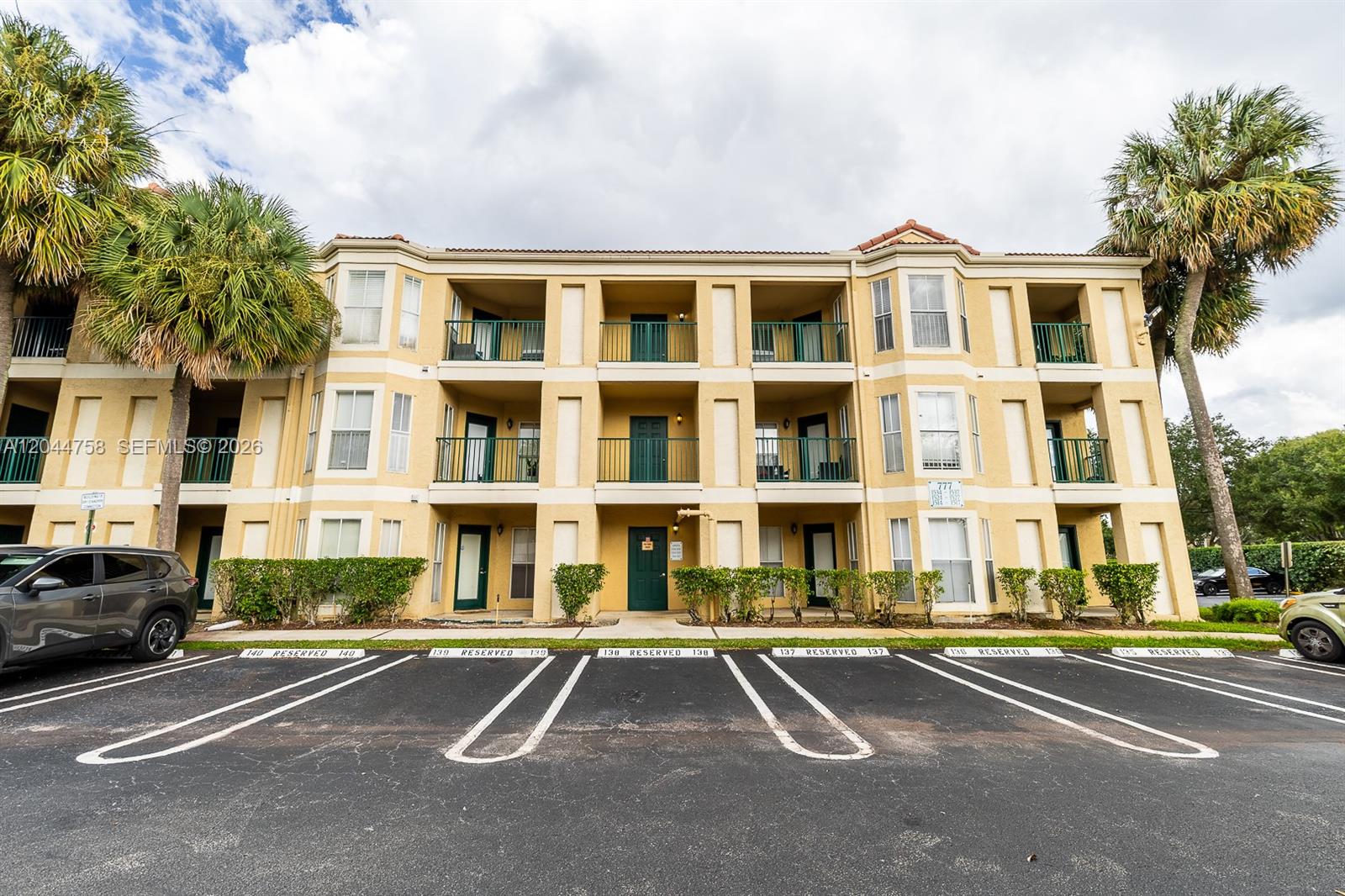 A three-story apartment building features yellow stucco walls, green balconies, and reserved parking spaces — CORAL SPRINGS, FL