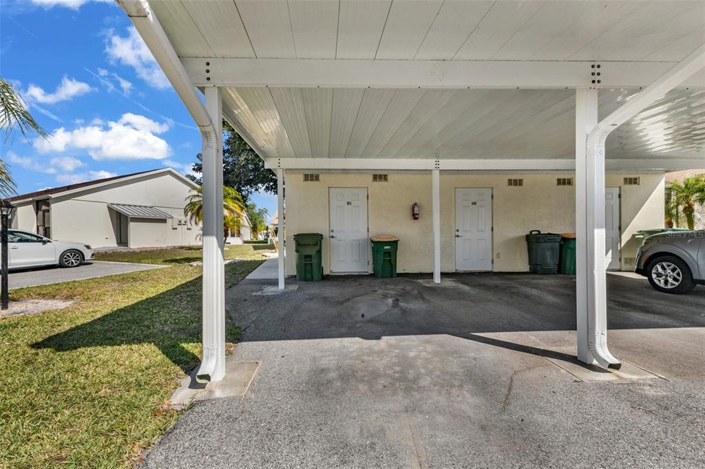A covered carport area features two white doors, green trash bins, and concrete flooring — 6796 GASPARILLA PINES BOULEVARD UNIT 105, ENGLEWOOD, FL