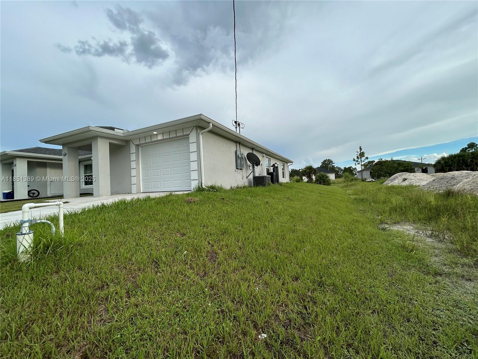 The exterior of a light-colored house features a garage, a side yard, and utility equipment — 1110 Augusta St E # 1110, Lehigh Acres FL 33974, LEHIGH ACRES, FL