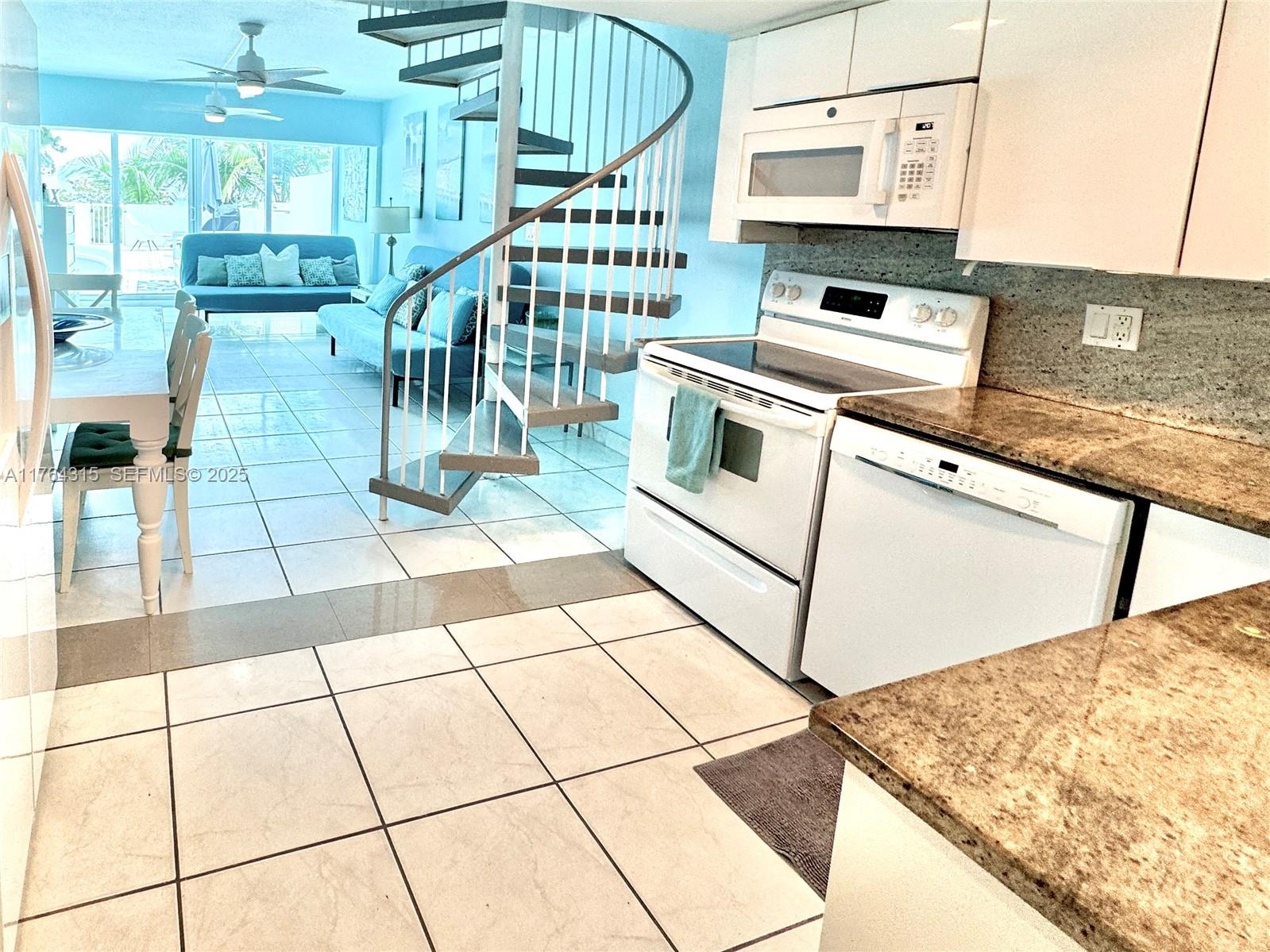 A kitchen with white appliances and granite countertops overlooks a living area with spiral stairs — 5445 Collins Ave # 11, Miami Beach FL 33140, MIAMI BEACH, FL