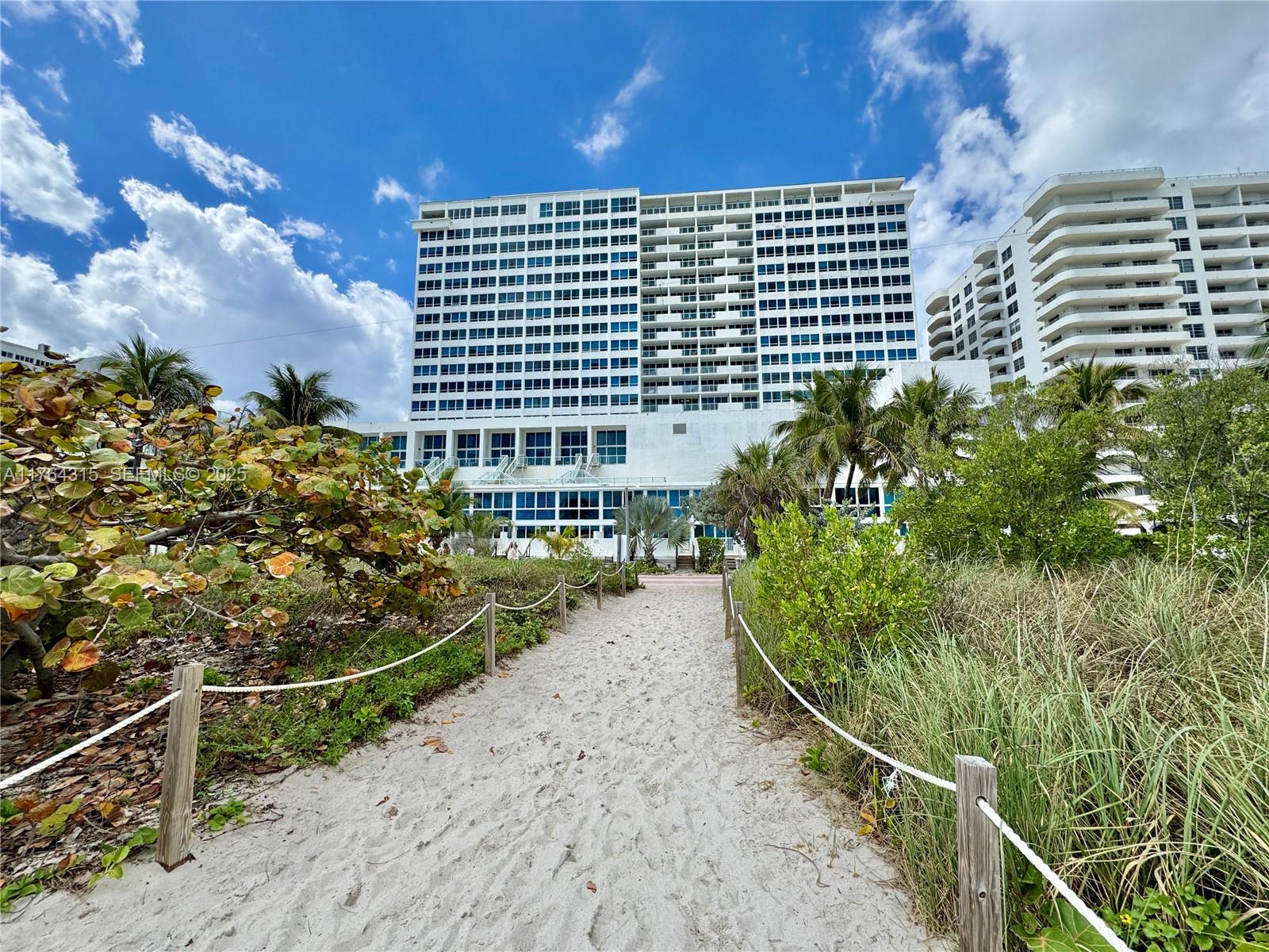 A sandy path leads toward a large white high-rise building surrounded by tropical vegetation — 5445 Collins Ave # 11, Miami Beach FL 33140, MIAMI BEACH, FL