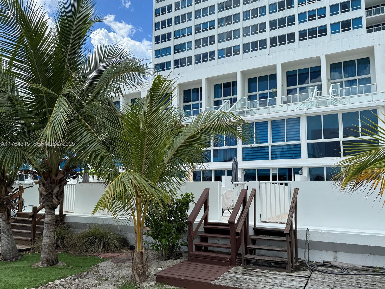A white multi-story building features balconies and wooden stairs leading to a patio area — 5445 Collins Ave # 11, Miami Beach FL 33140, MIAMI BEACH, FL