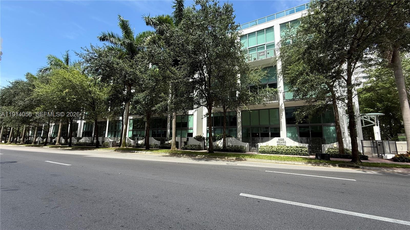A multi-story white building with large windows is partially obscured by trees along a road — 6000 Collins Ave # 305, Miami Beach FL 33140, MIAMI BEACH, FL