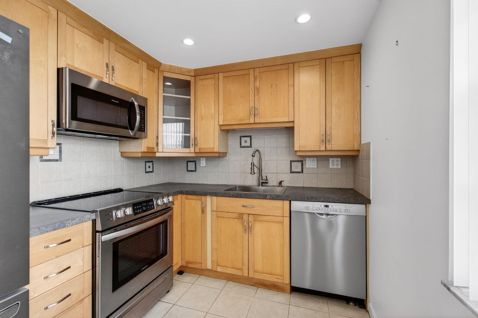 A kitchen features light wood cabinets, stainless steel appliances, a dark countertop, and tiled backsplash — 167 Normandy # D, Delray Beach FL 33484, DELRAY BEACH, FL