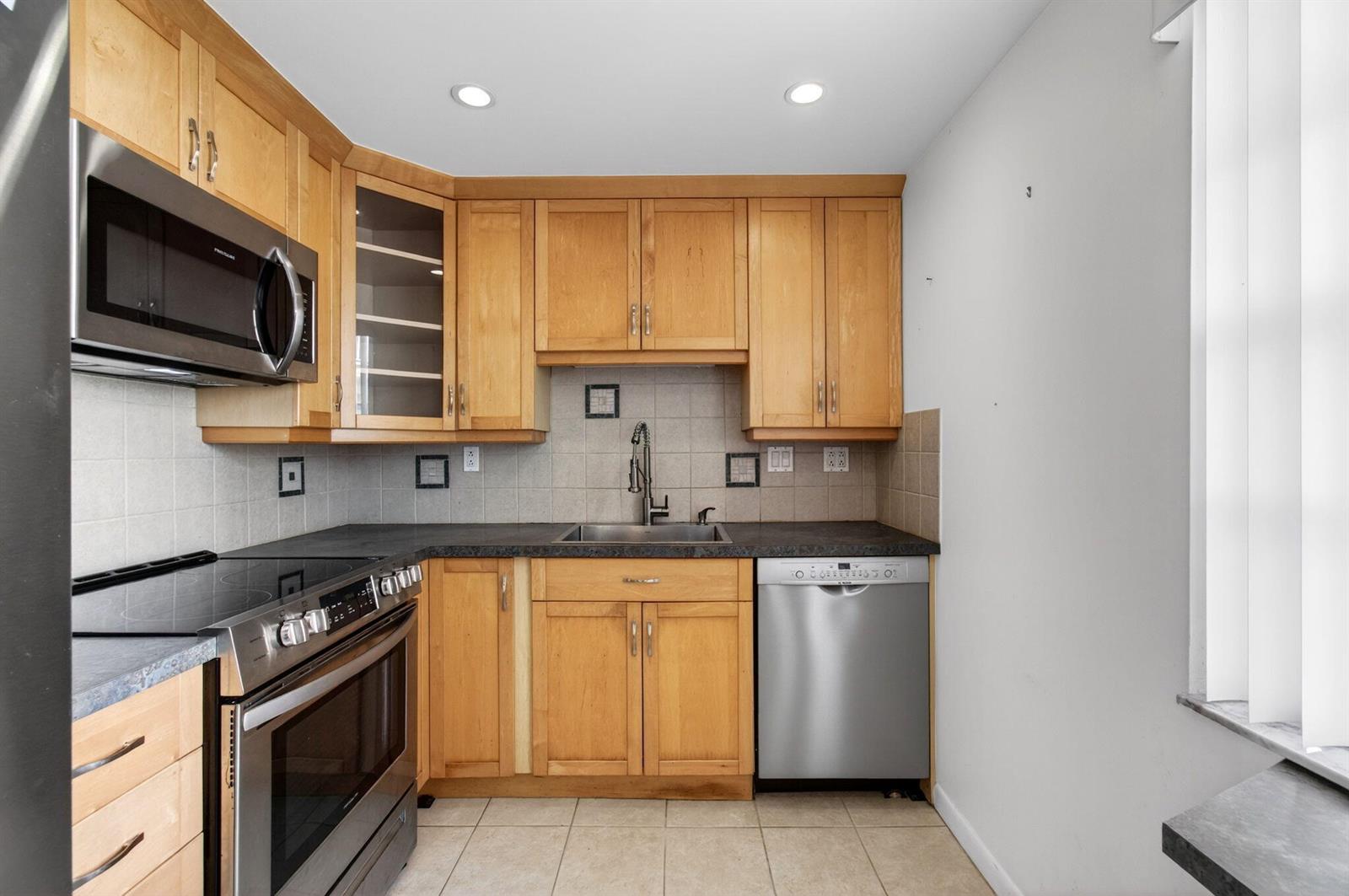 A kitchen features light wood cabinets, stainless steel appliances, dark countertops, and tiled flooring — 167 Normandy # D, Delray Beach FL 33484, DELRAY BEACH, FL