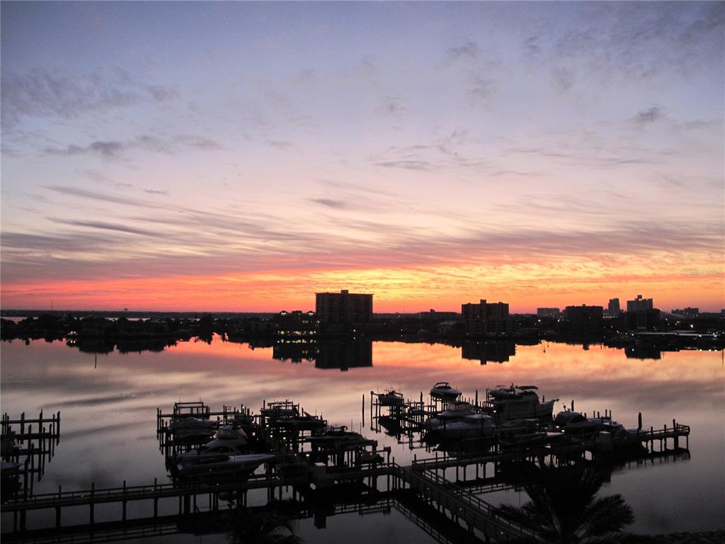 A sunset view over a marina with docked boats and distant buildings reflected in water — 521 MANDALAY AVENUE UNIT 704, CLEARWATER BEACH, FL