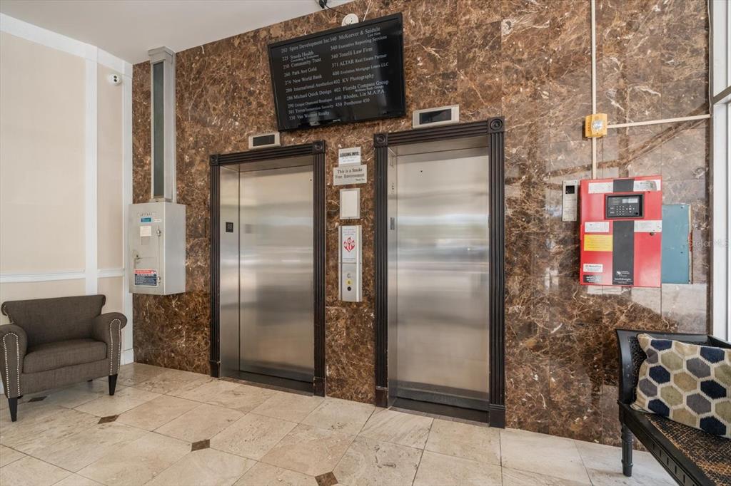 An elevator lobby features two stainless steel elevators set against a brown marble wall — 157 E NEW ENGLAND AVENUE UNIT 302, WINTER PARK, FL