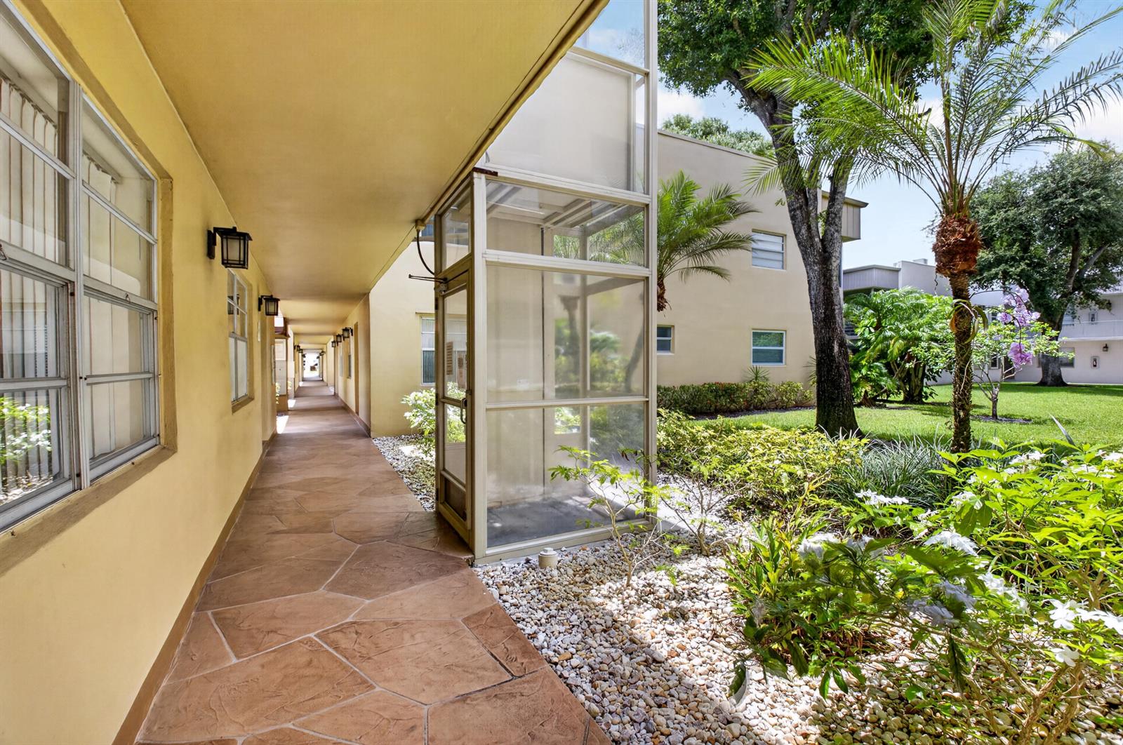 An outdoor walkway with stone flooring leads past a glass-enclosed elevator shaft and landscaping — 506 E Capri Road # 506, Delray Beach FL 33484, DELRAY BEACH, FL