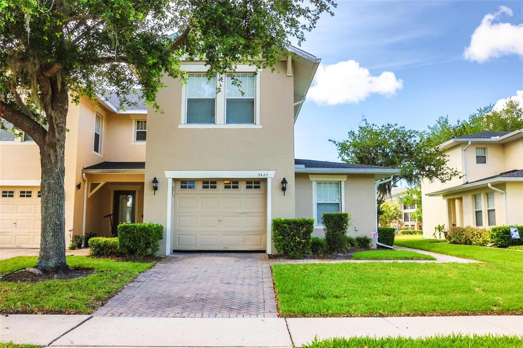 A two-story tan house with a brick driveway, a garage, and a large front tree — 5428 CARAMELLA DRIVE, ORLANDO, FL