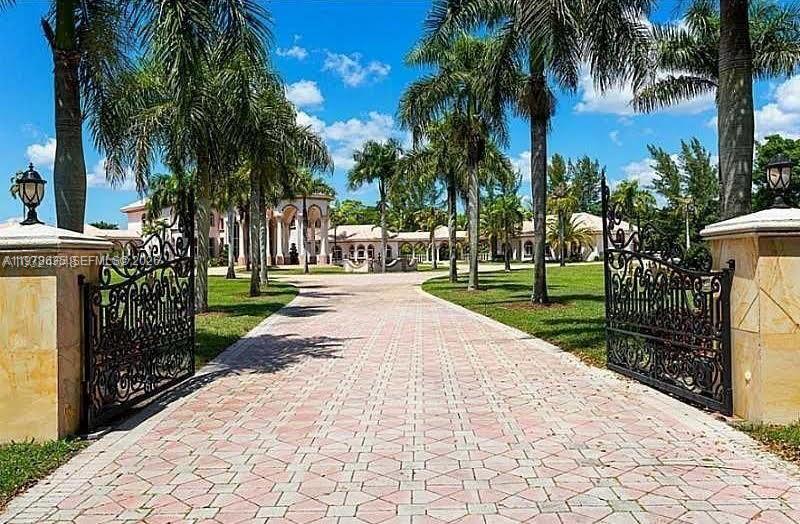 A paved driveway leads through an ornate iron gate toward a large estate with palms — 21579 Cartagena Dr, Boca Raton FL 33428, BOCA RATON, FL