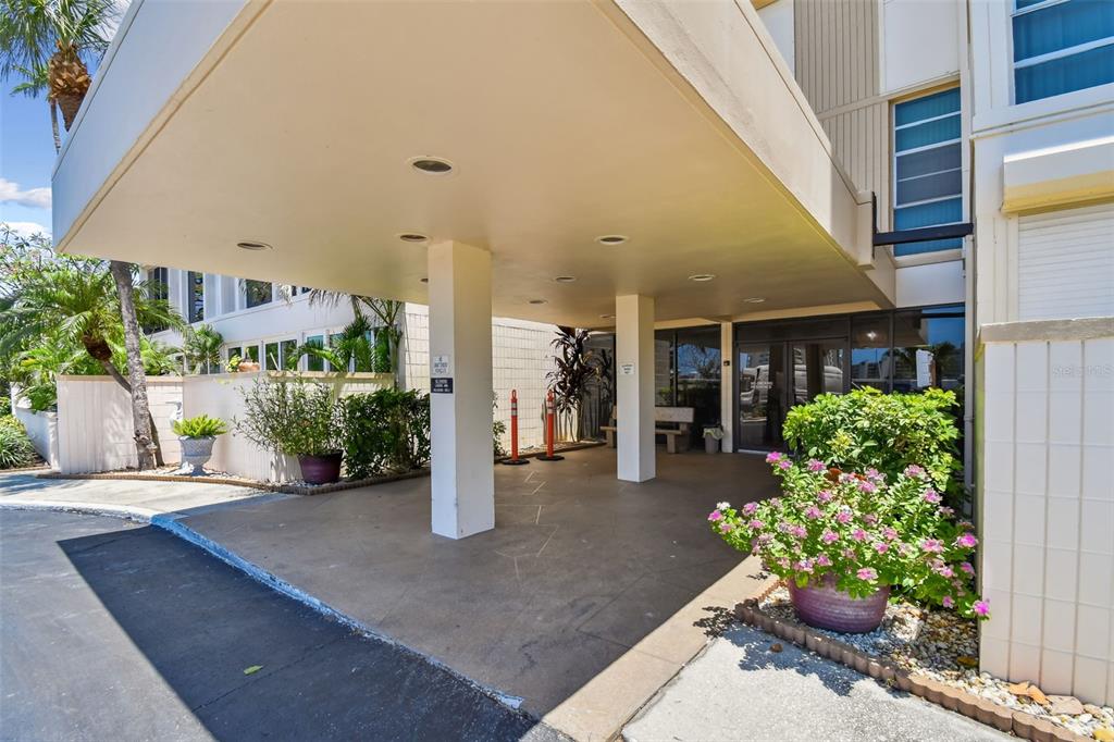 A covered building entrance features a concrete driveway, support pillars, and potted flowering plants — 7400 SUN ISLAND DRIVE S UNIT 411, SOUTH PASADENA, FL
