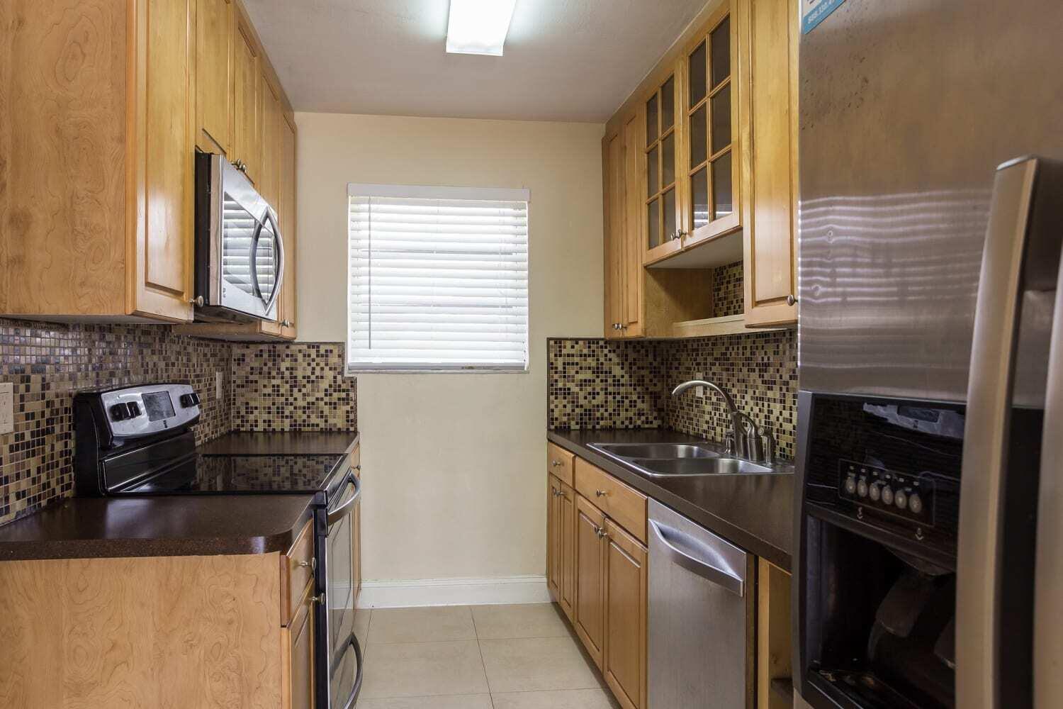 A galley kitchen features light wood cabinets, dark countertops, a mosaic tile backsplash, and stainless appliances — MIAMI, FL