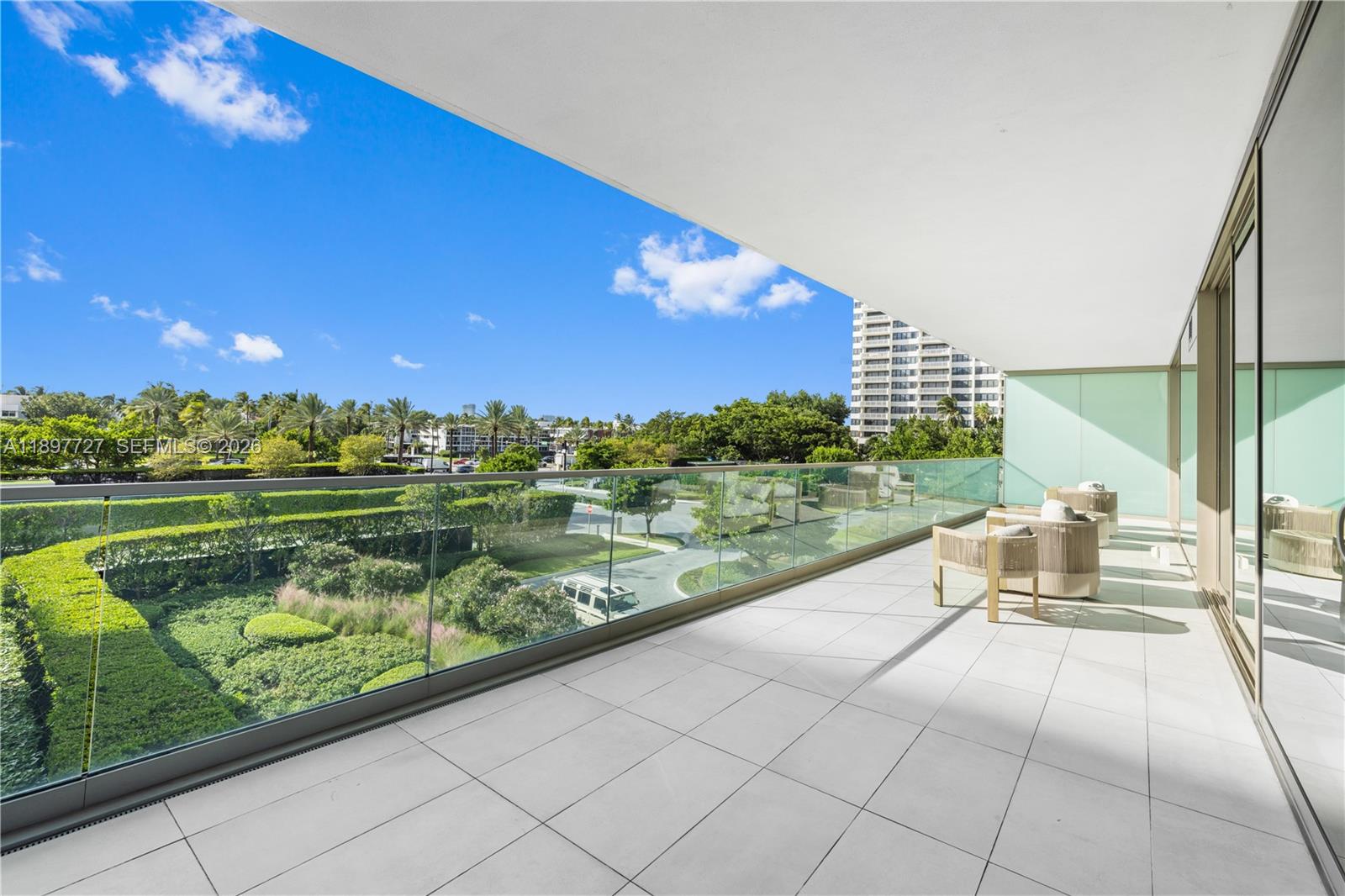 A spacious balcony features white tile flooring, glass railings, and outdoor seating overlooking lush landscaping — 10201 Collins Ave # 311, Bal Harbour FL 33154, BAL HARBOUR, FL