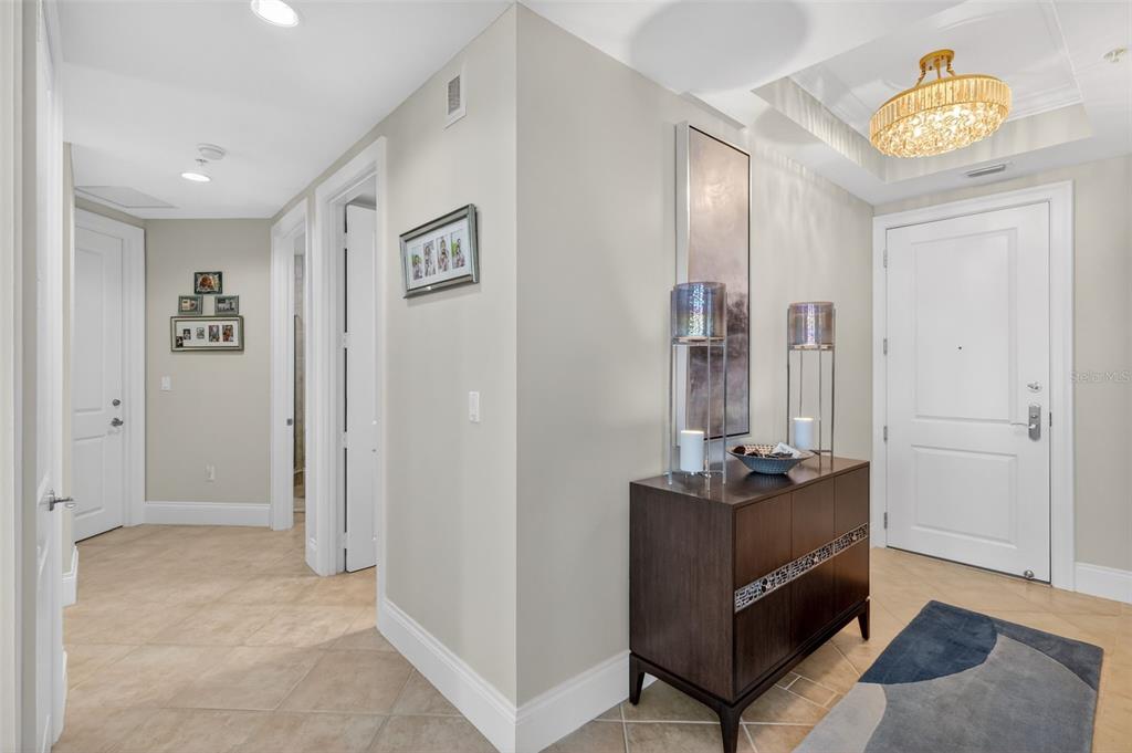 A bright entryway features a dark wood console table, a crystal chandelier, and tiled flooring — 130 RIVIERA DUNES WAY UNIT 506, PALMETTO, FL