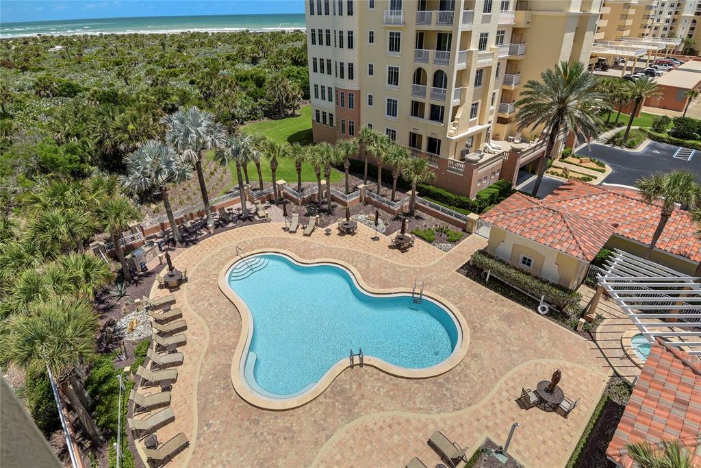 An aerial view shows a swimming pool with lounge chairs and palm trees near a building — 261 MINORCA BEACH WAY UNIT 606, NEW SMYRNA BEACH, FL