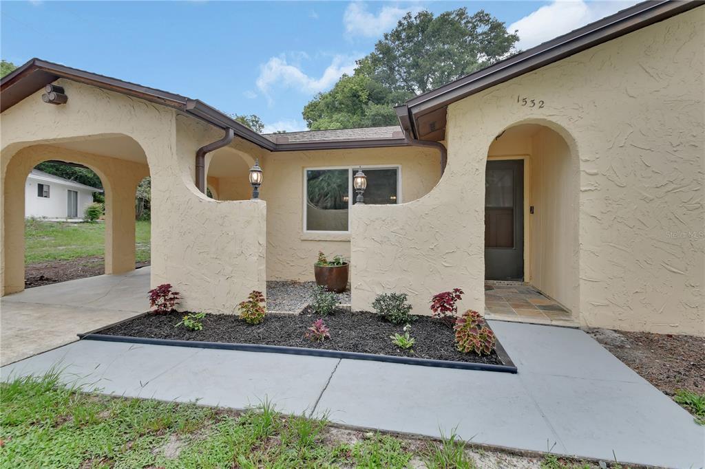 The exterior of a beige stucco house features a covered entryway, carport, and landscaped garden — 1532 N NORMANDY BOULEVARD, DELTONA, FL