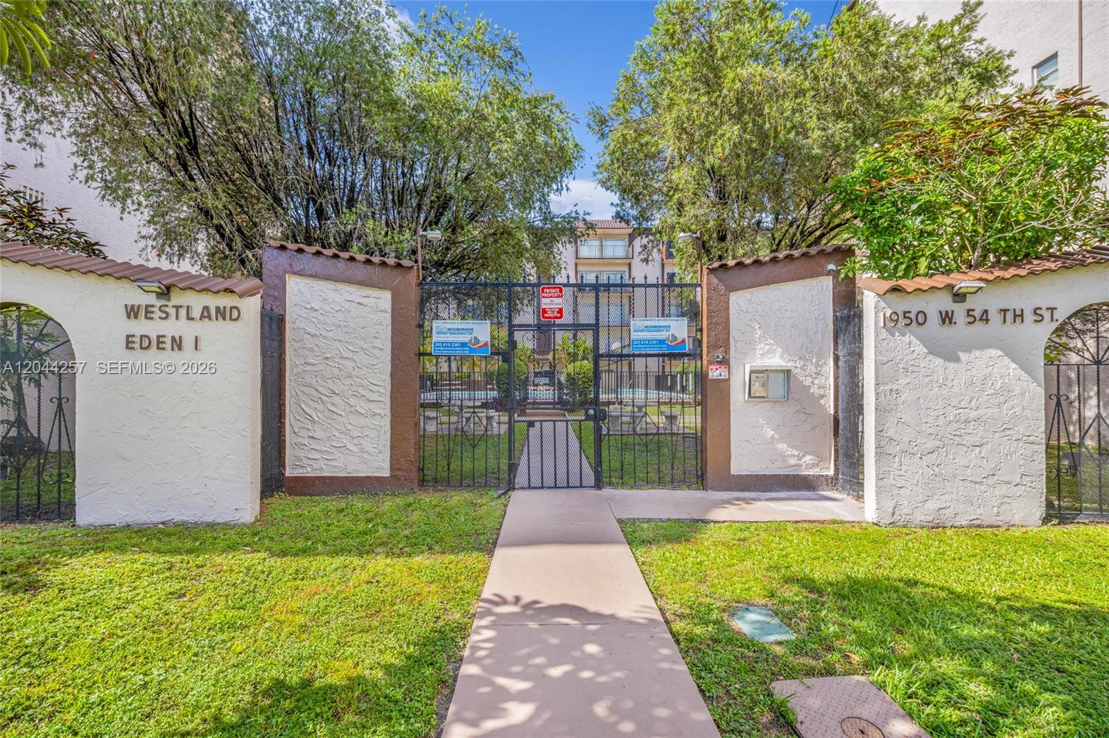 A gated entrance with a concrete walkway leads to a residential building with mature trees — 1950 W 54th St # 206, Hialeah FL 33012, HIALEAH, FL