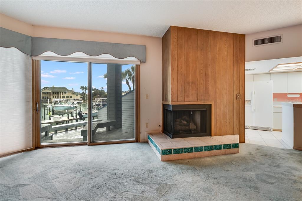 A living room features a wood-paneled corner fireplace, gray carpeting, and sliding glass doors overlooking a marina — 900 PINELLAS BAYWAY S UNIT 109, TIERRA VERDE, FL