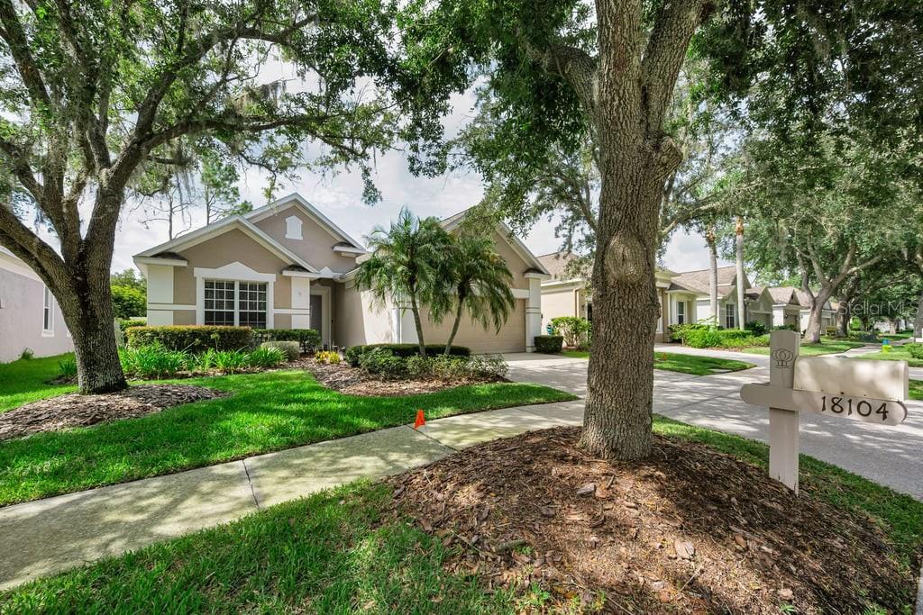 A single-story suburban house with a tan facade, a two-car garage, and mature oak trees — 18104 HAMDEN PARK WAY, TAMPA, FL
