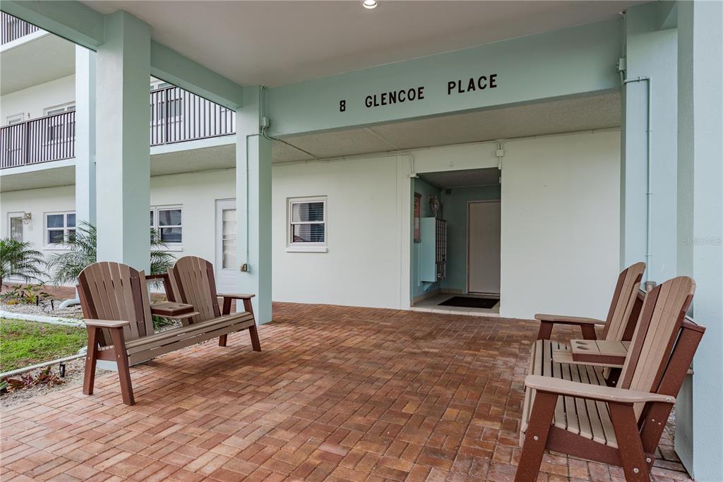 A covered brick patio features two wooden benches in front of a building entrance — 8 GLENCOE PLACE UNIT 104, DUNEDIN, FL