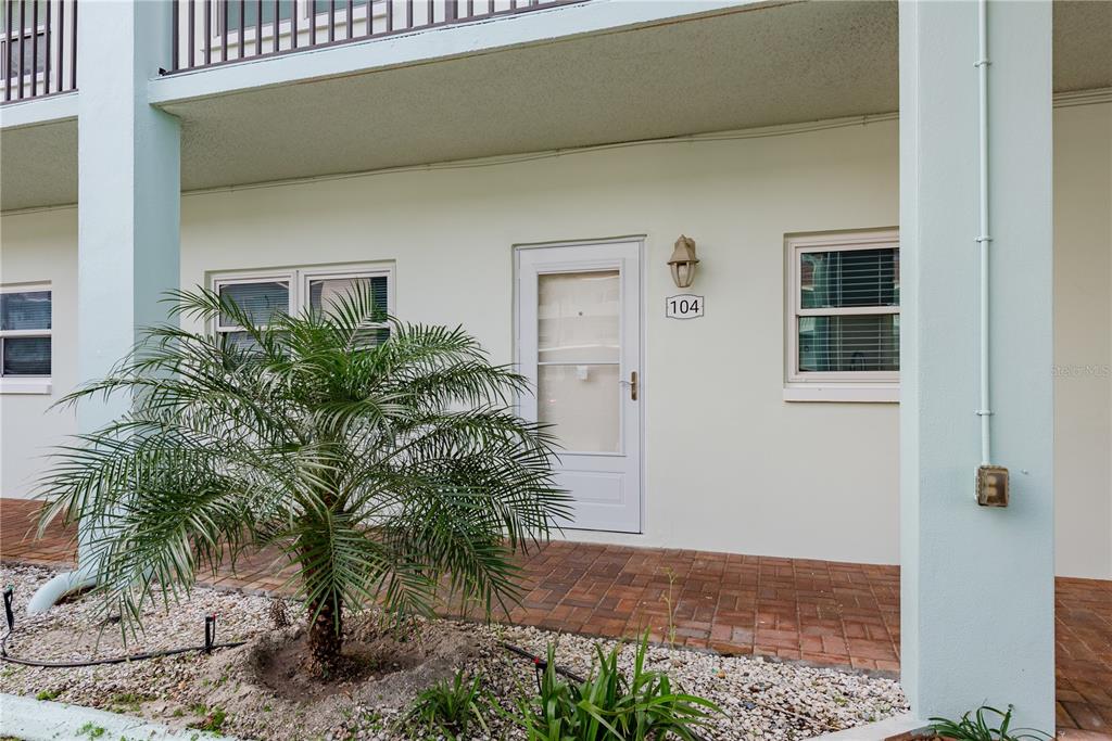 The exterior of a ground-floor apartment unit features a brick patio and a palm tree — 8 GLENCOE PLACE UNIT 104, DUNEDIN, FL