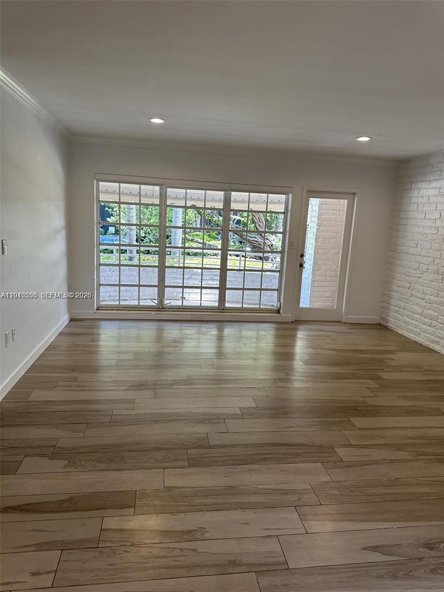 An empty room features light wood-look flooring, white walls, a brick accent wall, and large windows — BAL HARBOUR, FL