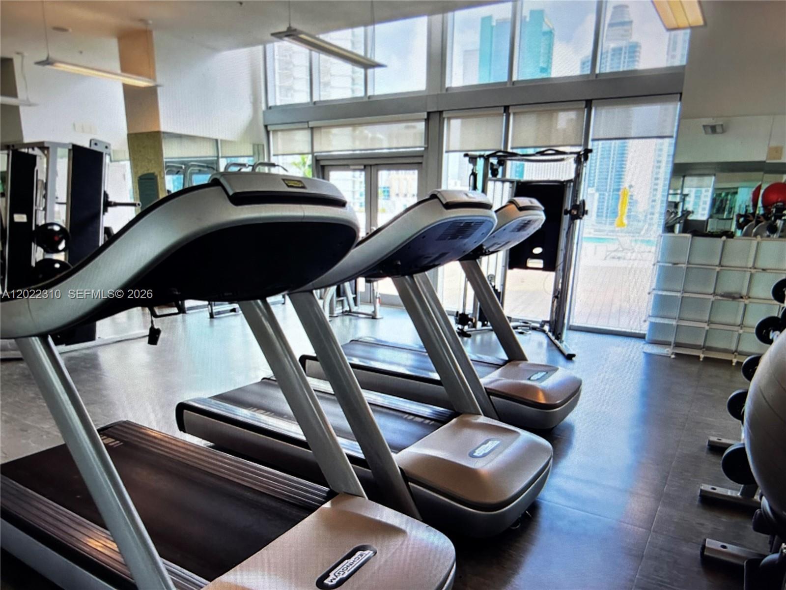A gym features three treadmills, a cable machine, and floor-to-ceiling windows overlooking city buildings — 92 SW 3rd St # 3703, Miami FL 33130, MIAMI, FL