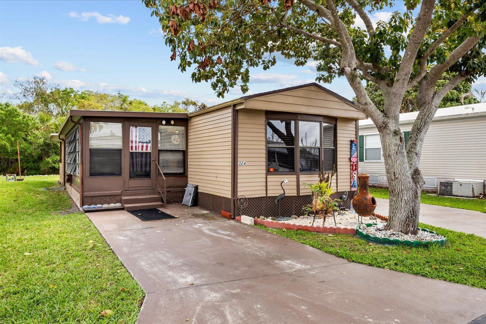 A tan mobile home with a brown roof and a concrete driveway under a tree — 324 Mockingbird Avenue, Fort Pierce FL 34982, FORT PIERCE, FL