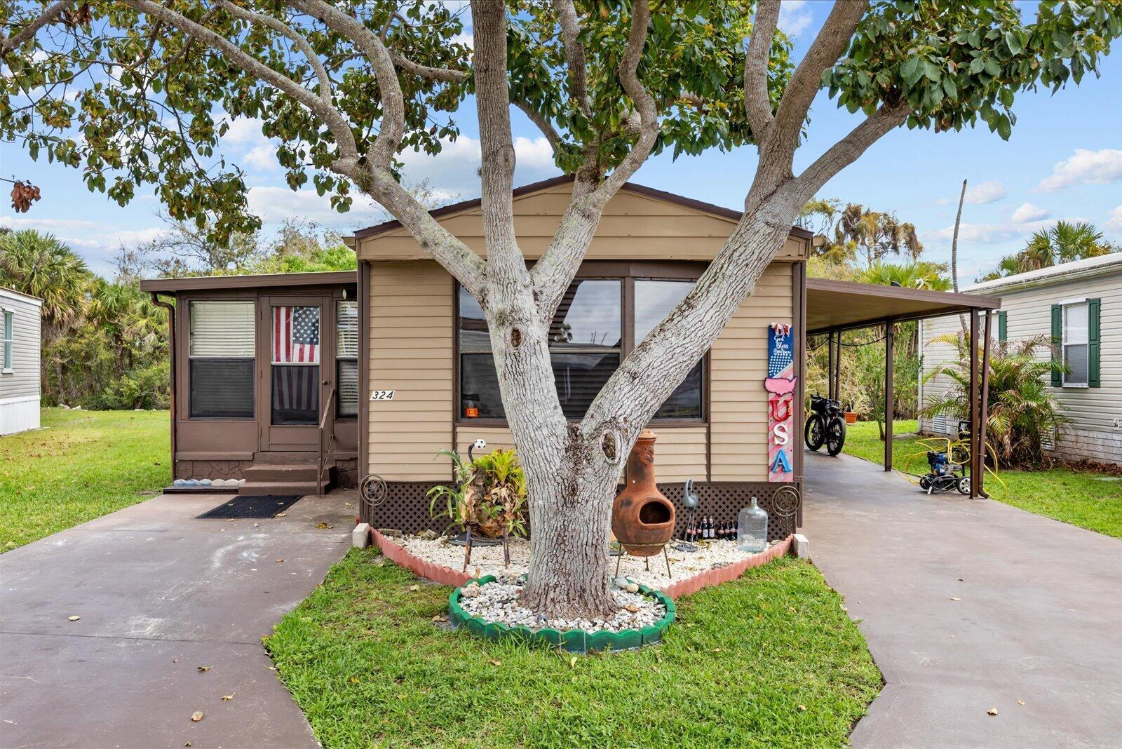 A single-story mobile home with tan siding, a front tree, and a covered carport — 324 Mockingbird Avenue, Fort Pierce FL 34982, FORT PIERCE, FL