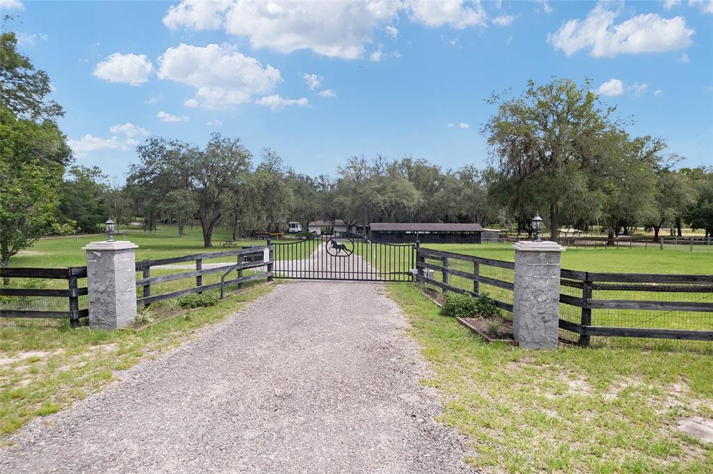 A gravel driveway leads to a gated entrance with stone pillars and wooden fencing — 10871 SE COUNTY ROAD 336, MORRISTON, FL