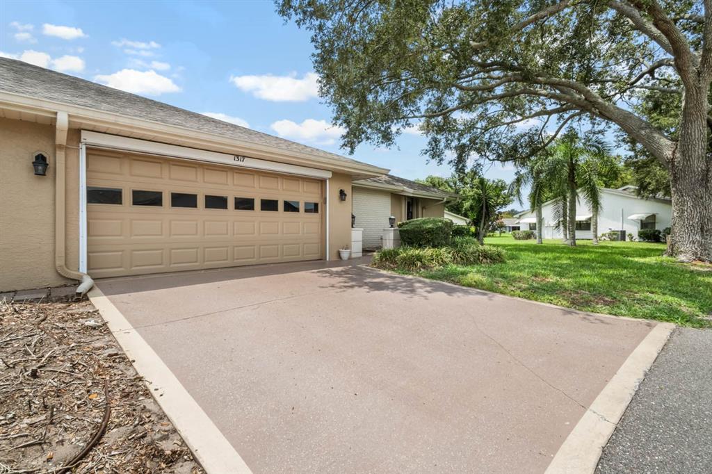 A tan house exterior features a two-car garage, a concrete driveway, and a large oak tree — 1317 LELAND DRIVE, SUN CITY CENTER, FL