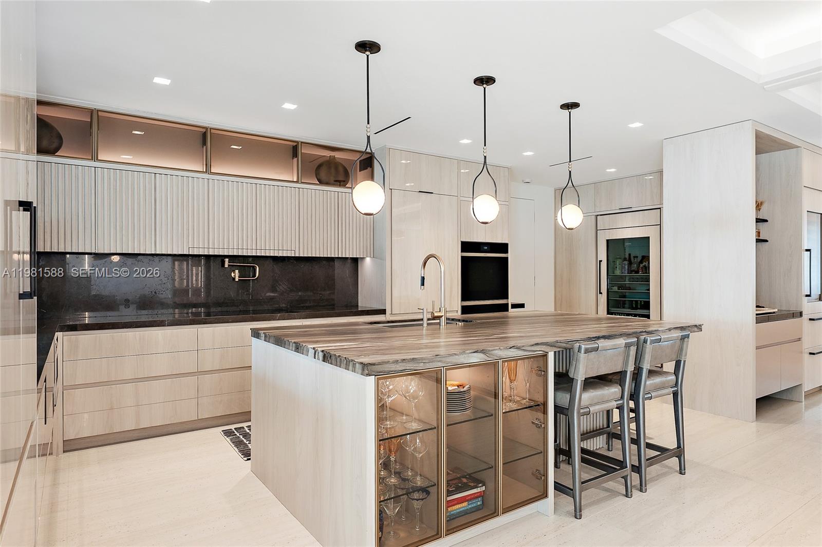 A modern kitchen features light wood cabinetry, a stone island with integrated glass shelving, and pendant lighting — 3501 Captains Way, Jupiter FL 33477, JUPITER, FL