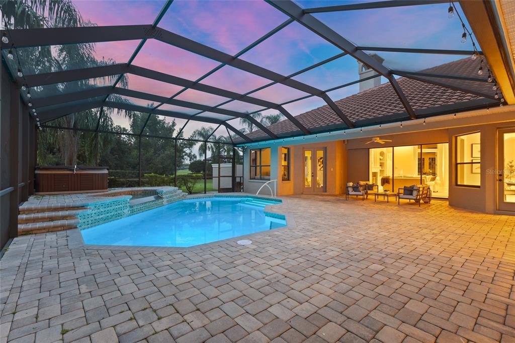 A screened-in patio features a swimming pool, a hot tub, and paved flooring at dusk — 7808 DERBY CT, LAKEWOOD RANCH, FL