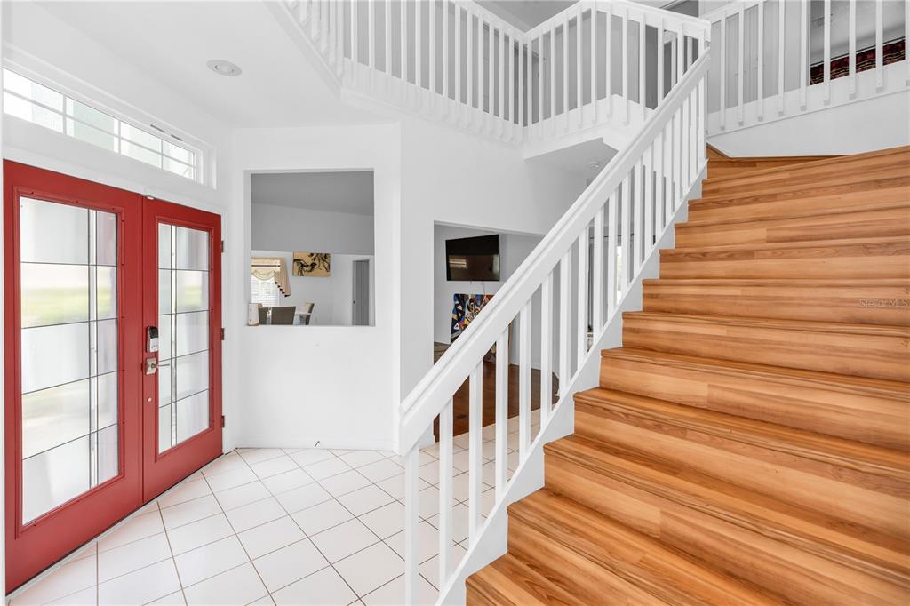 A bright foyer features white tile floors, a red double door, and a wooden staircase — 219 STRATHMORE CIRCLE, KISSIMMEE, FL