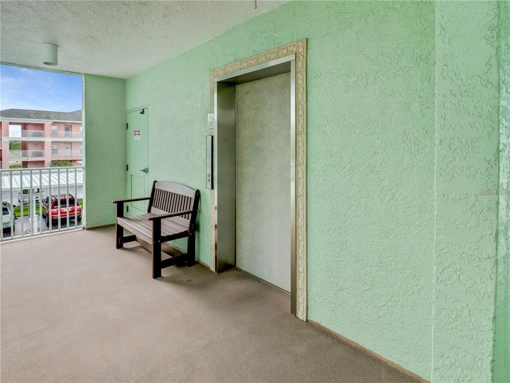 An outdoor apartment hallway features a light green wall, an elevator, and a wooden bench — 2121 HERON LAKE DRIVE UNIT 204, PUNTA GORDA, FL