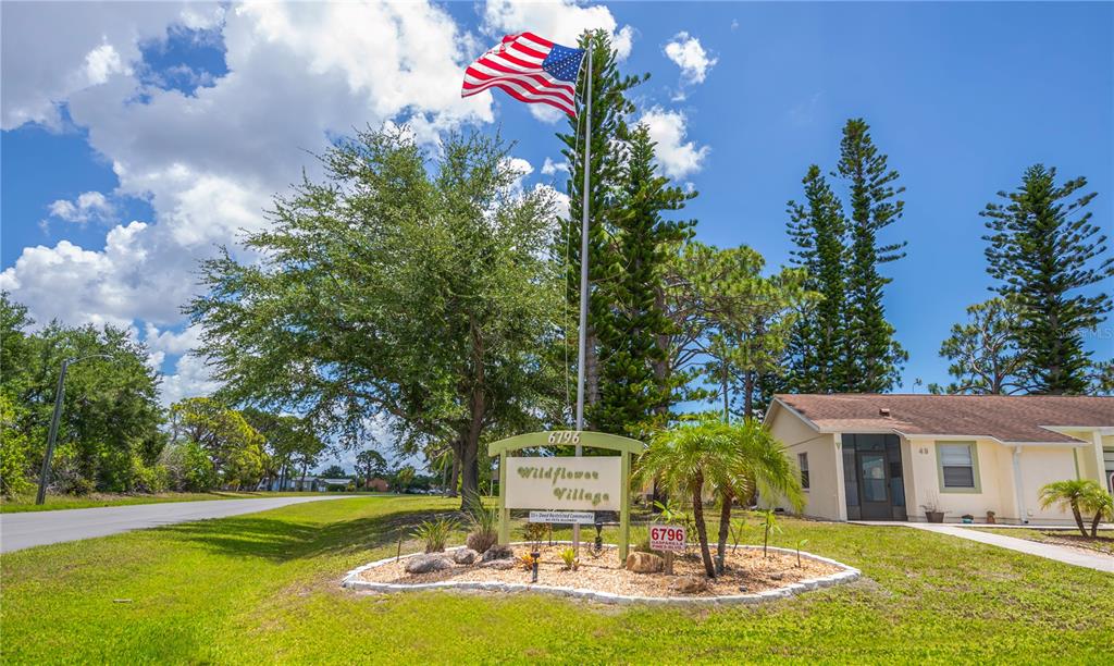 A sign for Wildflower Village stands in a landscaped island near a residential building — 6796 GASPARILLA PINES BOULEVARD UNIT 10, ENGLEWOOD, FL